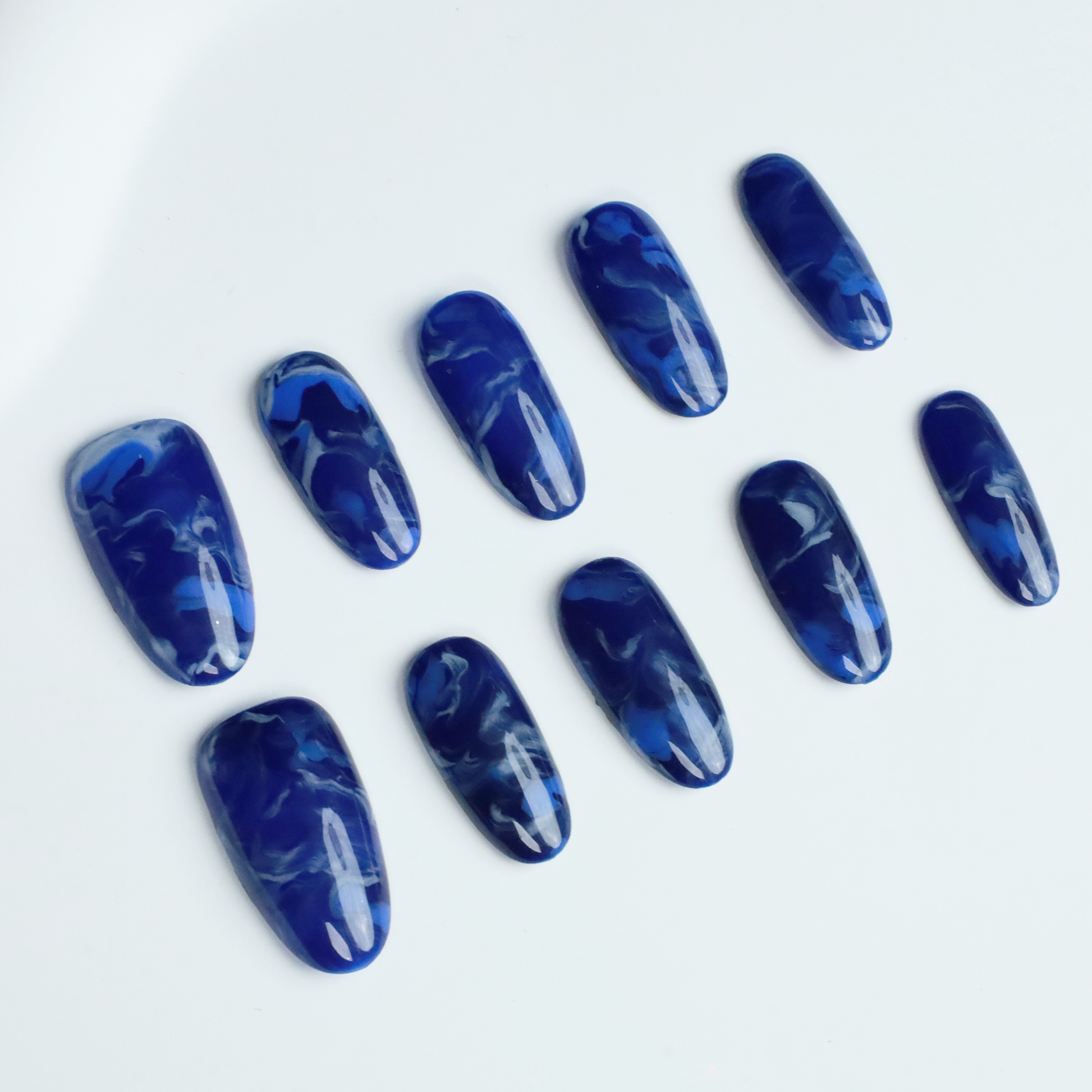 Hands display the Elegant Blue Marble Press-On Nails from We Do Nails, featuring glossy long oval shapes with a blue marble effect. A black ribbed sweater against a black background highlights these striking nails.