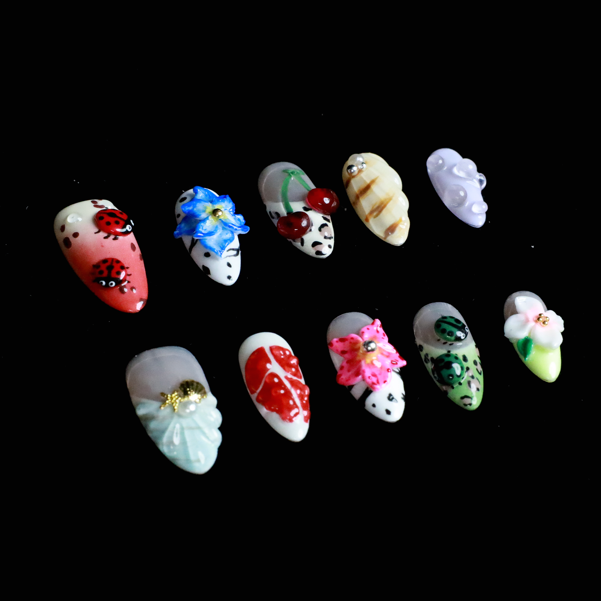 The Garden Blooms Press-On Nails by We Do Nails feature a whimsical garden theme with long, decorative nails, including a blue flower, cherries, red and white patterns, pearls, and stripes. Set against a black background, the hand is partially covered by a black sleeve.