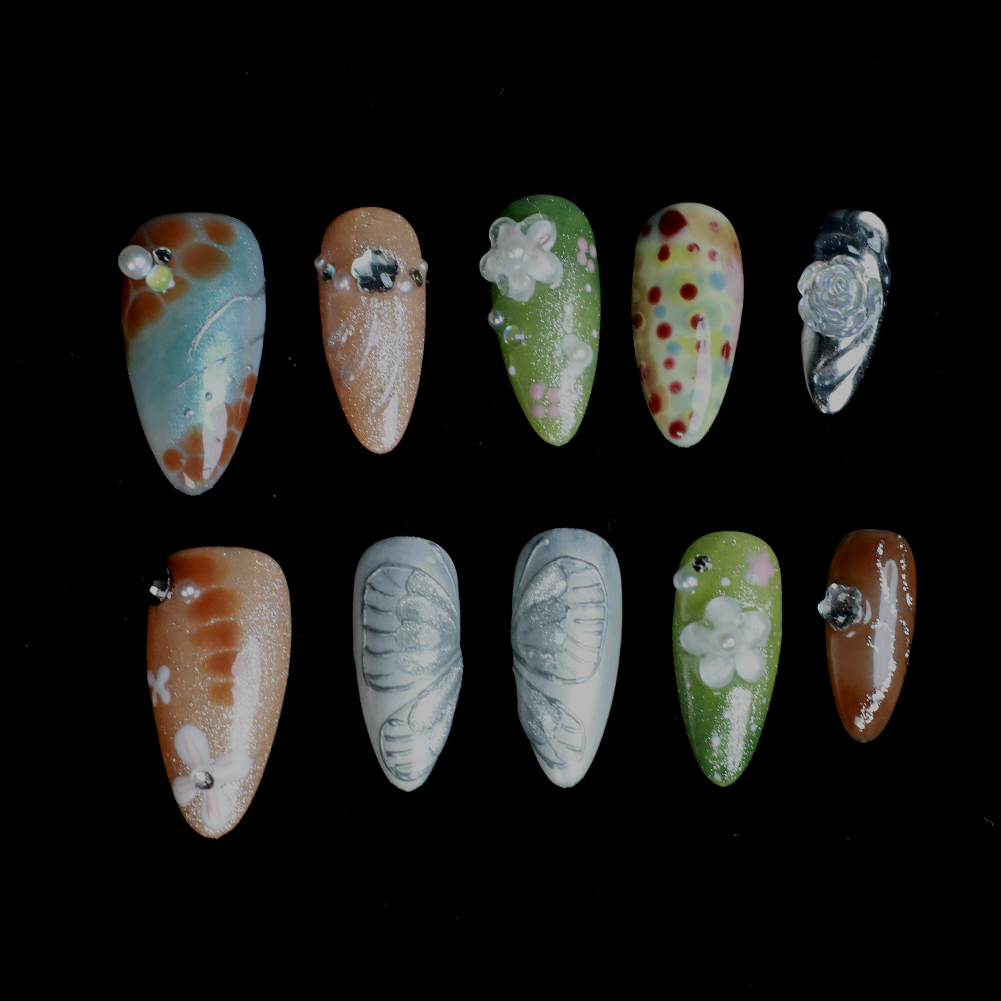 A hand showcases the Fantasy Garden Press-On Nails by We Do Nails against a black background. The nails feature designs of a koi fish with a pearl, peach with rhinestones, 3D green flowers, green with red floral dots, and metallic silver with a crystal accent.