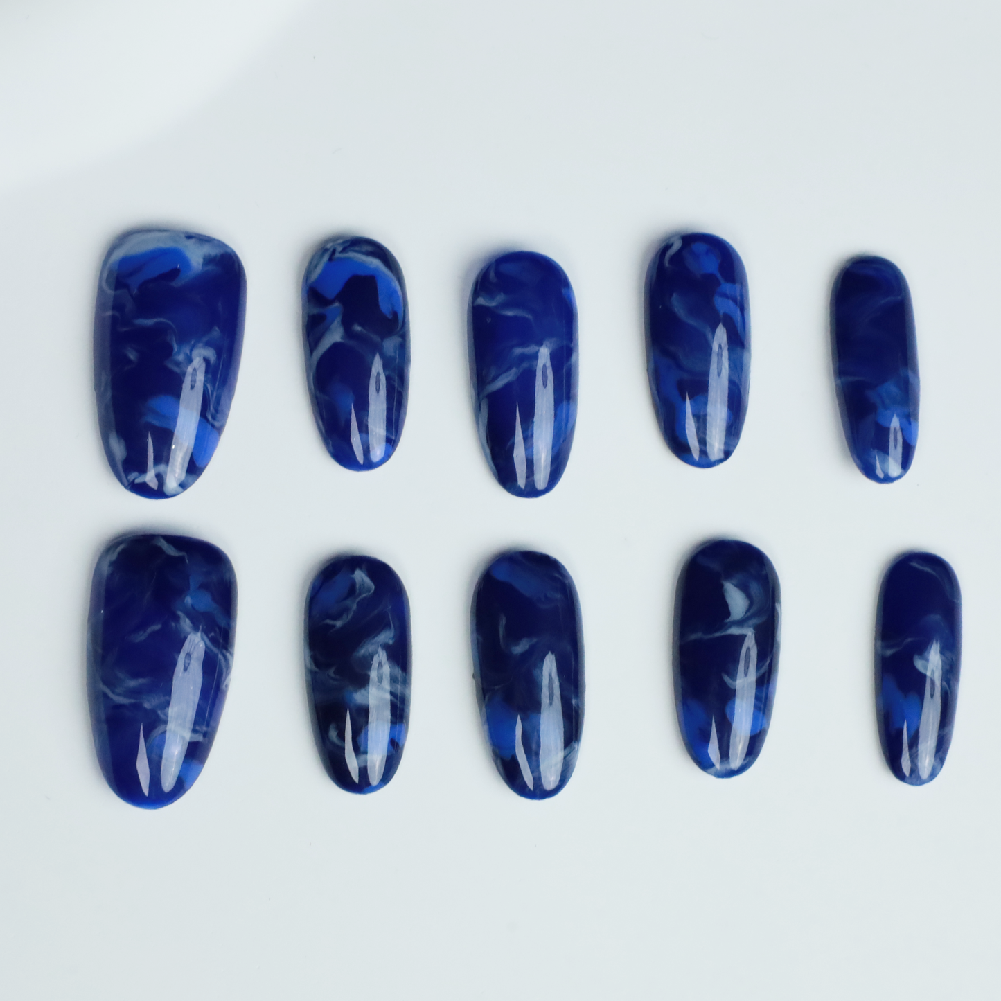 Hands display the Elegant Blue Marble Press-On Nails from We Do Nails, featuring glossy long oval shapes with a blue marble effect. A black ribbed sweater against a black background highlights these striking nails.