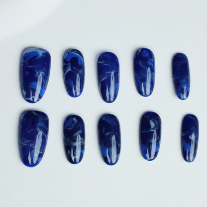 Hands display the Elegant Blue Marble Press-On Nails from We Do Nails, featuring glossy long oval shapes with a blue marble effect. A black ribbed sweater against a black background highlights these striking nails.