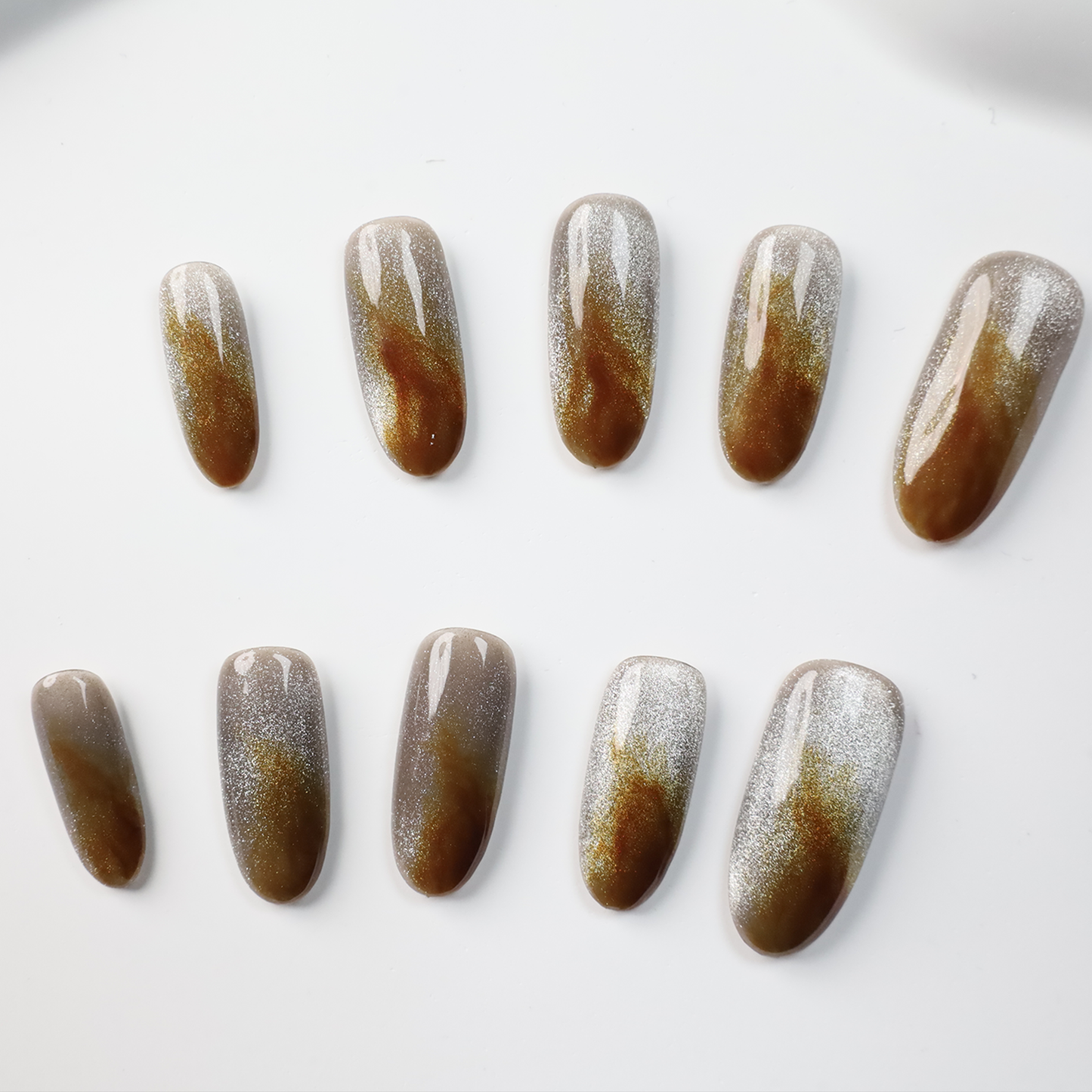 A hand with We Do Nails Elegant Brown Marble Press-On Nails, featuring a subtle metallic chrome accent, holds a simple white mug against a black backdrop.