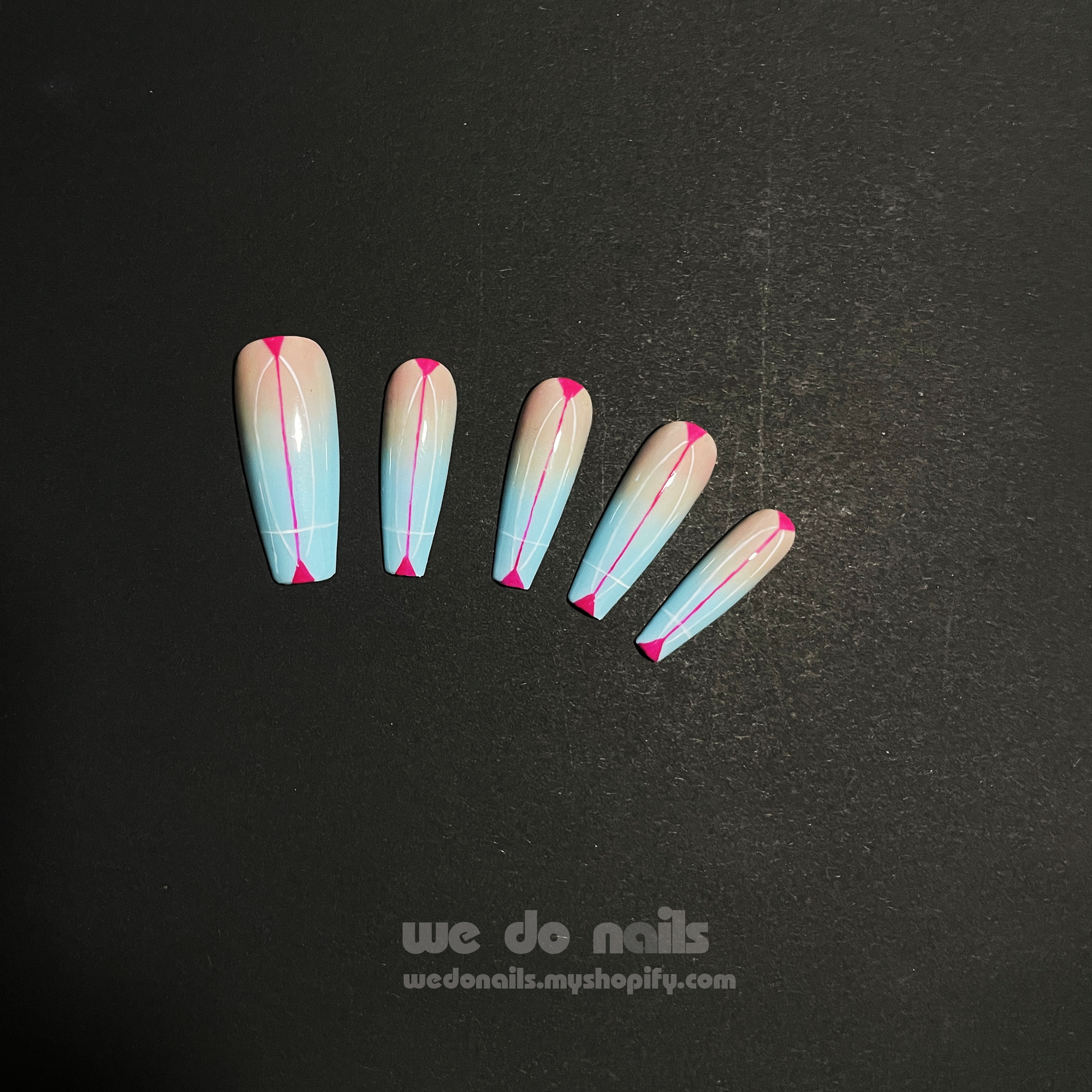 Close-up of hands with long, stiletto-shaped Sky Dream Press-On Nails by We Do Nails, featuring a glossy pastel blue with pink tips. A silver ring with a pearl graces one finger against a dark, striped fabric backdrop that enhances the elegant style.