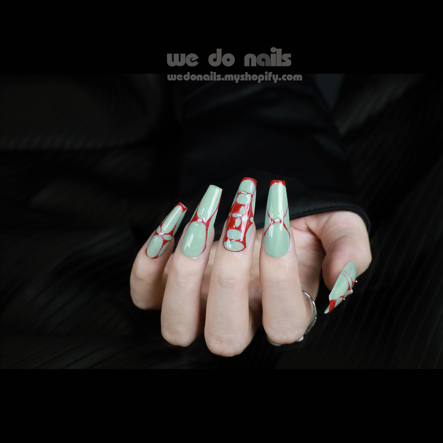 A partially covered hand in a black sleeve displays Passionate Love Press-On Nails from We Do Nails, featuring long, pointed tips with intricate red lines and dots over mint green polish. These reusable nails allow for customizable designs and a unique style each time, resting on a dark textured surface.
