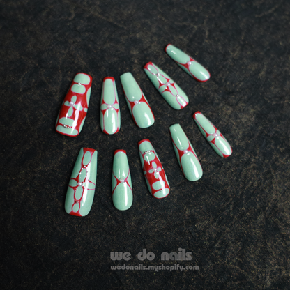 A partially covered hand in a black sleeve displays Passionate Love Press-On Nails from We Do Nails, featuring long, pointed tips with intricate red lines and dots over mint green polish. These reusable nails allow for customizable designs and a unique style each time, resting on a dark textured surface.